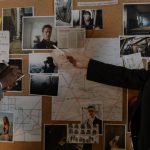 Dois investigadores analisam um painel com fotografias, mapas e anotações durante uma investigação criminal.