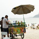 Vendedor de bebidas trabalha sob um guarda-sol em uma praia do Rio de Janeiro, com o mar ao fundo.