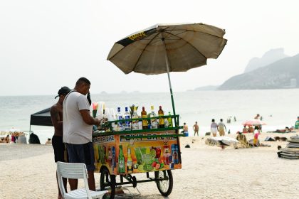 Vendedor de bebidas trabalha sob um guarda-sol em uma praia do Rio de Janeiro, com o mar ao fundo.