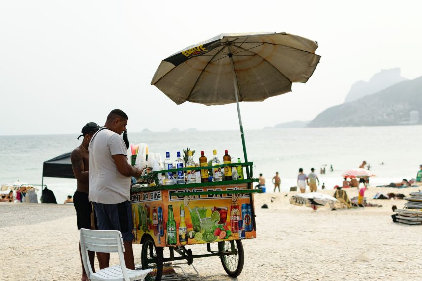 Vendedor de bebidas trabalha sob um guarda-sol em uma praia do Rio de Janeiro, com o mar ao fundo.