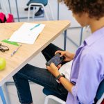 Criança joga em um celular sentada à carteira, com materiais escolares ao redor, durante uma aula.