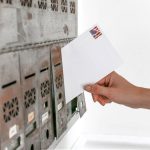 Mão inserindo um envelope selado em uma caixa de correio, representando o envio de um voto pelo correio.