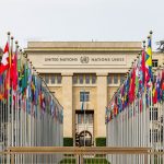 Vista do escritório das Nações Unidas em Genebra, com bandeiras de diversos países enfileiradas diante do edifício.