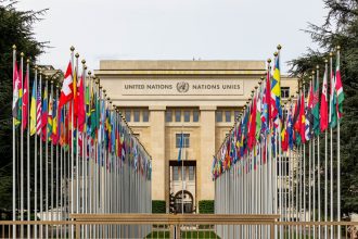 Vista do escritório das Nações Unidas em Genebra, com bandeiras de diversos países enfileiradas diante do edifício.