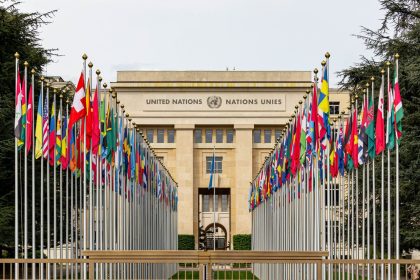 Vista do escritório das Nações Unidas em Genebra, com bandeiras de diversos países enfileiradas diante do edifício.