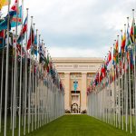 Bandeiras de diversos países diante da entrada da sede da ONU em Genebra, em um parque arborizado.