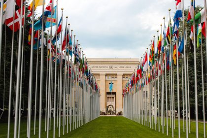 Bandeiras de diversos países diante da entrada da sede da ONU em Genebra, em um parque arborizado.