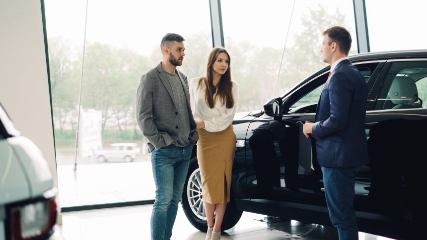 Casal conversa com um vendedor sobre a compra de um carro dentro de uma concessionária.