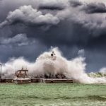 Ondas fortes quebram contra um píer e um farol sob um céu escuro e tempestuoso.