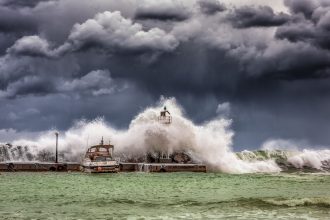 Ondas fortes quebram contra um píer e um farol sob um céu escuro e tempestuoso.